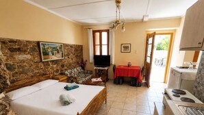 A bedroom with a bed, a TV, a dining table with chairs, a kitchen area with a stove, and a stone wall. - Elegant Studio in Chios Near the Beach and Port (Chios)