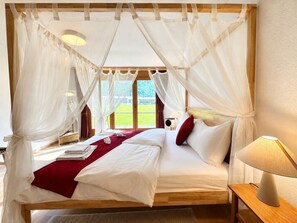 A four-poster bed with white linens and a red throw blanket, a bedside lamp, and a view of a green landscape through large windows. - Waterfall 3BR Garden Apt near Interlaken • AC by SwissDreamLiving (Meiringen)