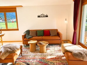 Un salon comprenant un canapé, deux petites tables en bois, un jeu d’échecs, un tapis à motifs et une vue sur un paysage verdoyant par la fenêtre.