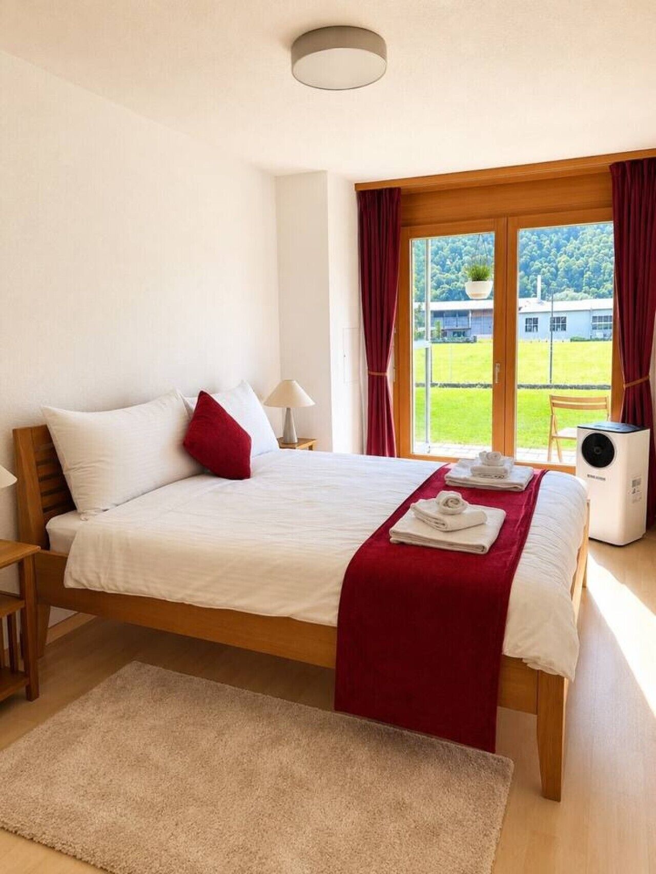 Een slaapkamer met een bed, nachtkastje, lamp, raam met gordijnen en uitzicht op groen.