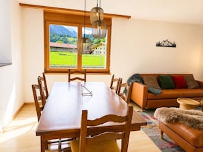 A dining area with a wooden table and chairs, a sofa, and a view of a green landscape through a large window.
