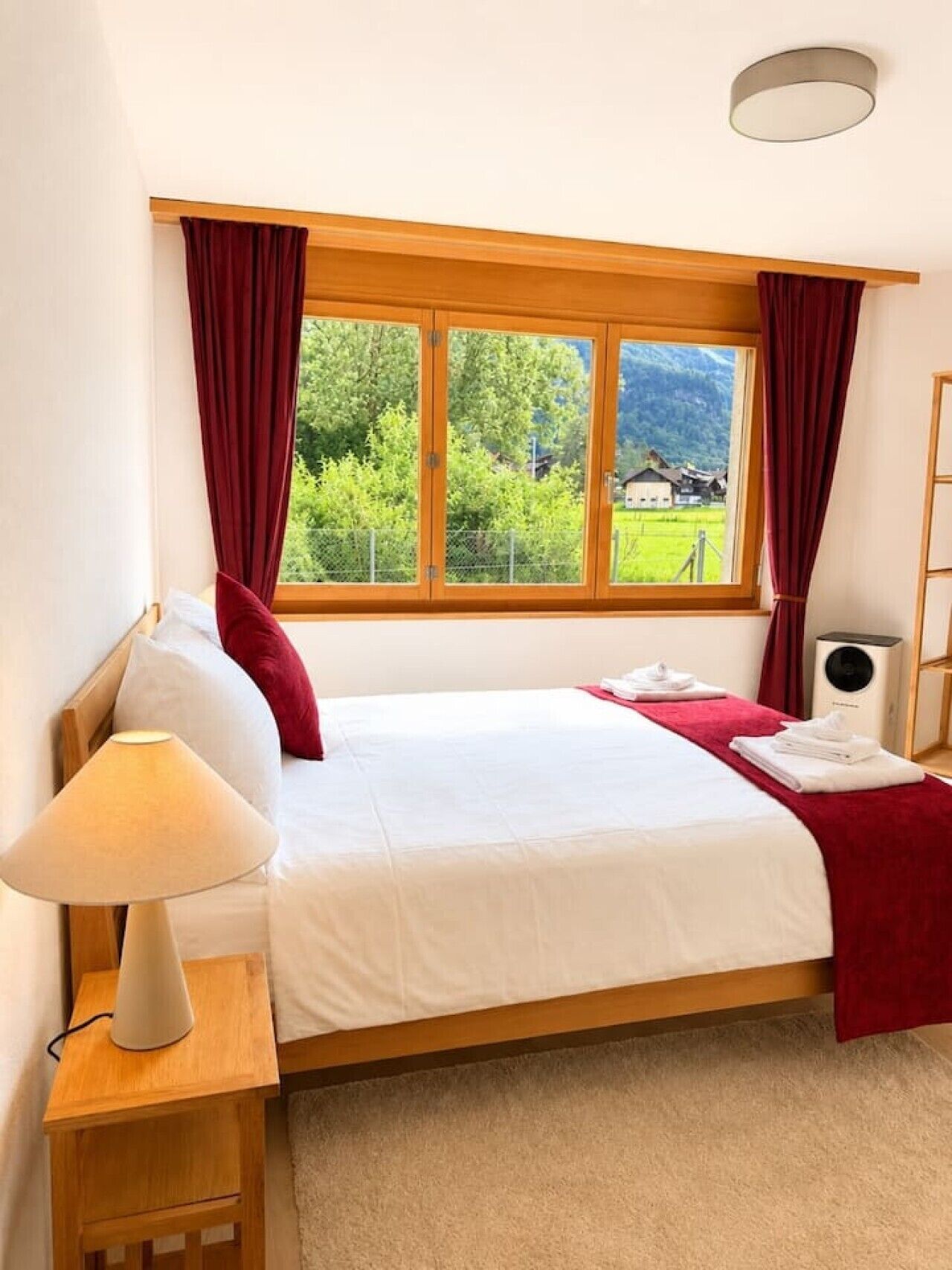 A bedroom with a bed, a bedside lamp, a window with curtains, and a view of greenery and a house.