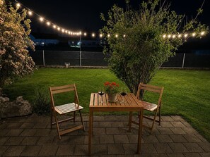 A patio with two wooden chairs and a table, a potted plant with red flowers, and string lights. - Waterfall 3BR Garden Apt near Interlaken • AC by SwissDreamLiving (Meiringen)