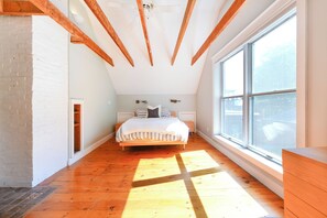 A bedroom with a wooden floor, a bed with a white and striped bedspread, a window with a view, and exposed wooden beams on the ceiling. - Upscale Historic West End Townhouse with Expansive Garden (Portland)