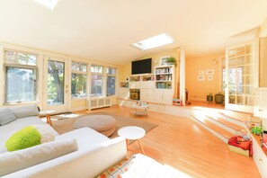 A spacious living room with a sofa, ottoman, and a television. There are bookshelves, a small table, and a staircase leading to another level. - Upscale Historic West End Townhouse with Expansive Garden (Portland)