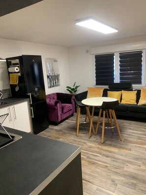 Un espace de vie compact comprenant un coin cuisine, un canapé violet, une table à manger avec des chaises et une fenêtre avec des stores.