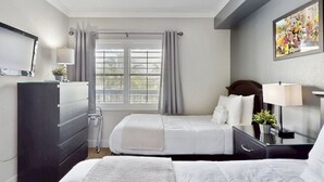 Habitación de hotel con dos camas, un escritorio, una silla, un aparador, una ventana con cortinas y un cuadro en la pared.