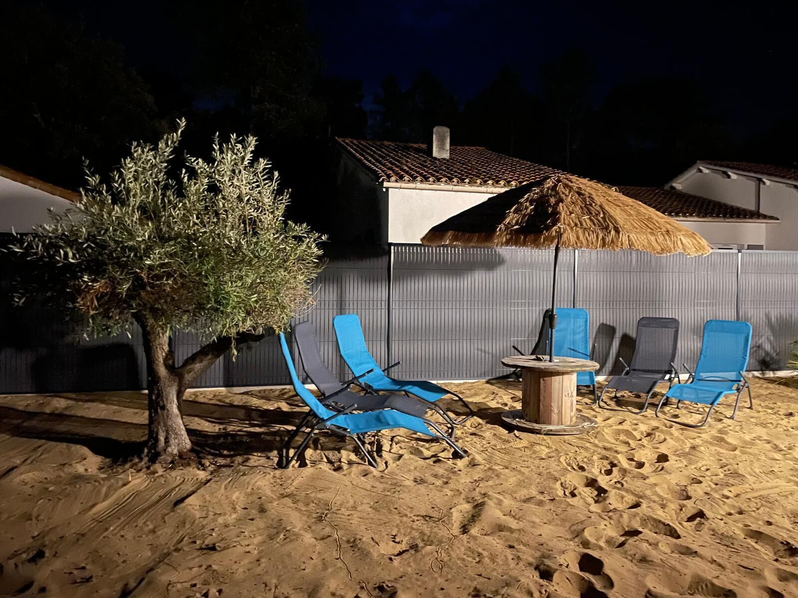 Un espacio al aire libre con arena, sillas reclinables azules, una mesita redonda y un paraguas de paja.