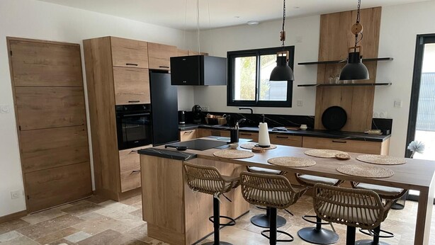 Eine moderne Küche mit Holzsäulen, einer zentralen Kochinsel mit Barhocker, einem schwarzen Kühlschrank und einem Fenster über der Spüle.