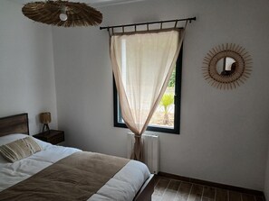 Ein Schlafzimmer mit einem Bett, einem Fenster mit durchsichtigen Vorhängen, einem Wandspiegel und einem Deckenventilator.