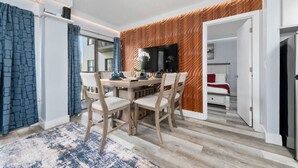 Una sala de estar moderna con comedor, un televisor de pantalla plana montado en una pared de madera, y un dormitorio visible con cama y almohada roja.