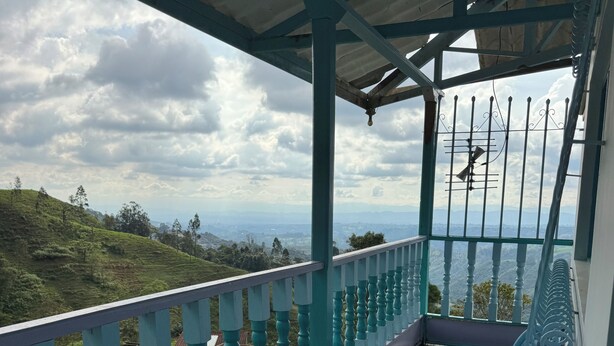 A covered balcony with a table and chairs, a railing, and a view of a hilly landscape.