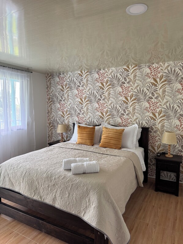 A neatly made bed with a patterned headboard, a bedside table with a lamp, and a window with sheer curtains.