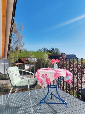 A balcony with a table and chairs, a satellite dish, and a view of a garden with blooming trees. - Cozy 3-bedroom vacation apartment with a private terrace and views of the mountains and lake (Hergensweiler)