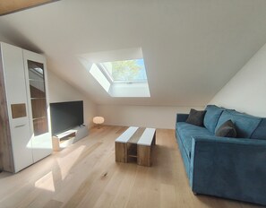 A modern loft bedroom with a skylight, a television, a blue sofa, and a wooden coffee table. - Cozy 3-bedroom vacation apartment with a private terrace and views of the mountains and lake (Hergensweiler)