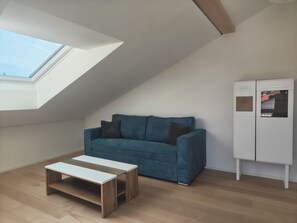 A room with a sloped ceiling, a blue sofa, a white cabinet, and a small wooden table. - Cozy 3-bedroom vacation apartment with a private terrace and views of the mountains and lake (Hergensweiler)