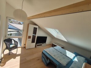 A room with a sloped ceiling, a television, a sofa, a chair, and a wardrobe. - Cozy 3-bedroom vacation apartment with a private terrace and views of the mountains and lake (Hergensweiler)