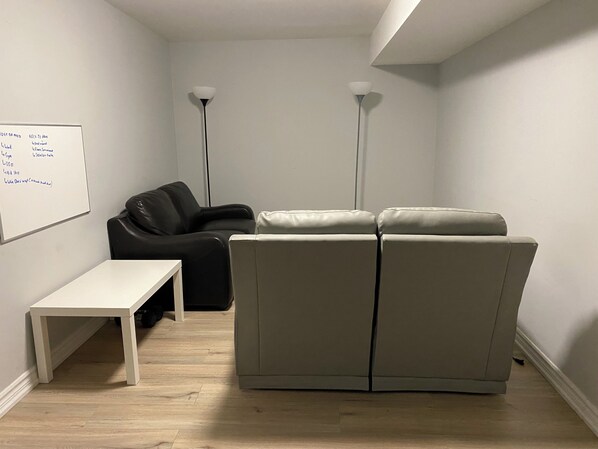 Ein Zimmer mit einer Tafel, einem schwarzen Stuhl, einem beigen Sofa, einem weißen Tisch und zwei Stehlampen.
