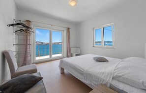 Une chambre à coucher avec un lit, une chaise, une étagère où sont suspendus des vêtements et une vue sur la mer grâce à de grandes fenêtres.