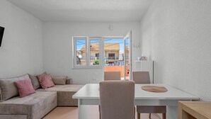 A modern living room with a white sofa, a dining table, and chairs. There is a window with a view of buildings outside. - Apartment right by the sea - 2 balconies, sea view of Krapanj (Brodarica)