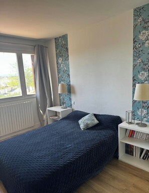 Ein Schlafzimmer mit einem Bett, einem Nachttisch, einer Lampe, einem Bücherregal und einem Fenster mit Vorhängen.