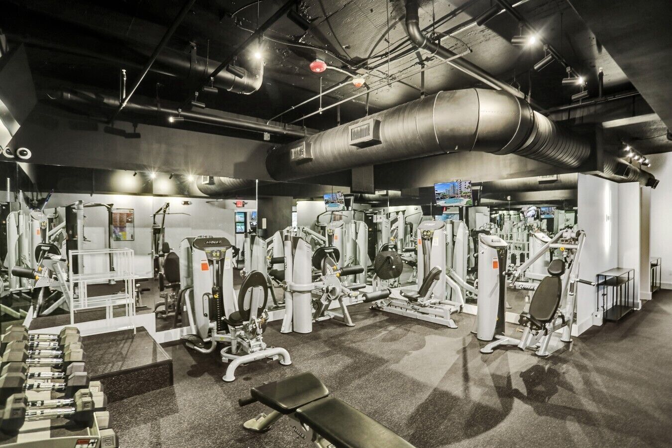 A gym with various exercise machines, a bench, and a large ventilation duct.