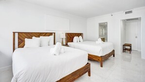 Una camera d'albergo con due letti, lenzuola bianche, poggiatesta in legno, un comodino con una lampada, uno sgabello di legno e un bagno con doccia in vetro.