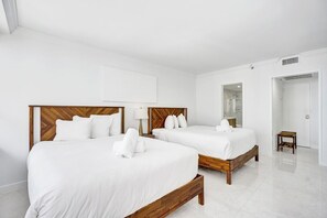 A hotel room with two beds, white linens, wooden headboards, a bedside table with a lamp, a small wooden stool, and a bathroom with a glass shower. - Spacious Ocean View Condo w/Resort Amenities 1108 (Miami Beach)