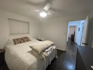 Un dormitorio con cama, ventilador de techo, mesita de noche y ventana con persianas, además de un pasillo que da a otra habitación.