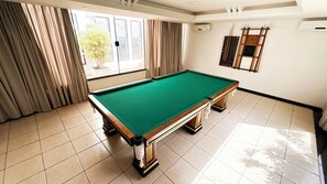 Uma mesa de bilhar em um espaço com janelas amplas, cortinas e piso ladrilhado.