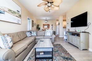 Ein Wohnzimmer mit Sofa, Couchtisch, Fernseher, Deckenventilator und einem Küchenbereich im Hintergrund.