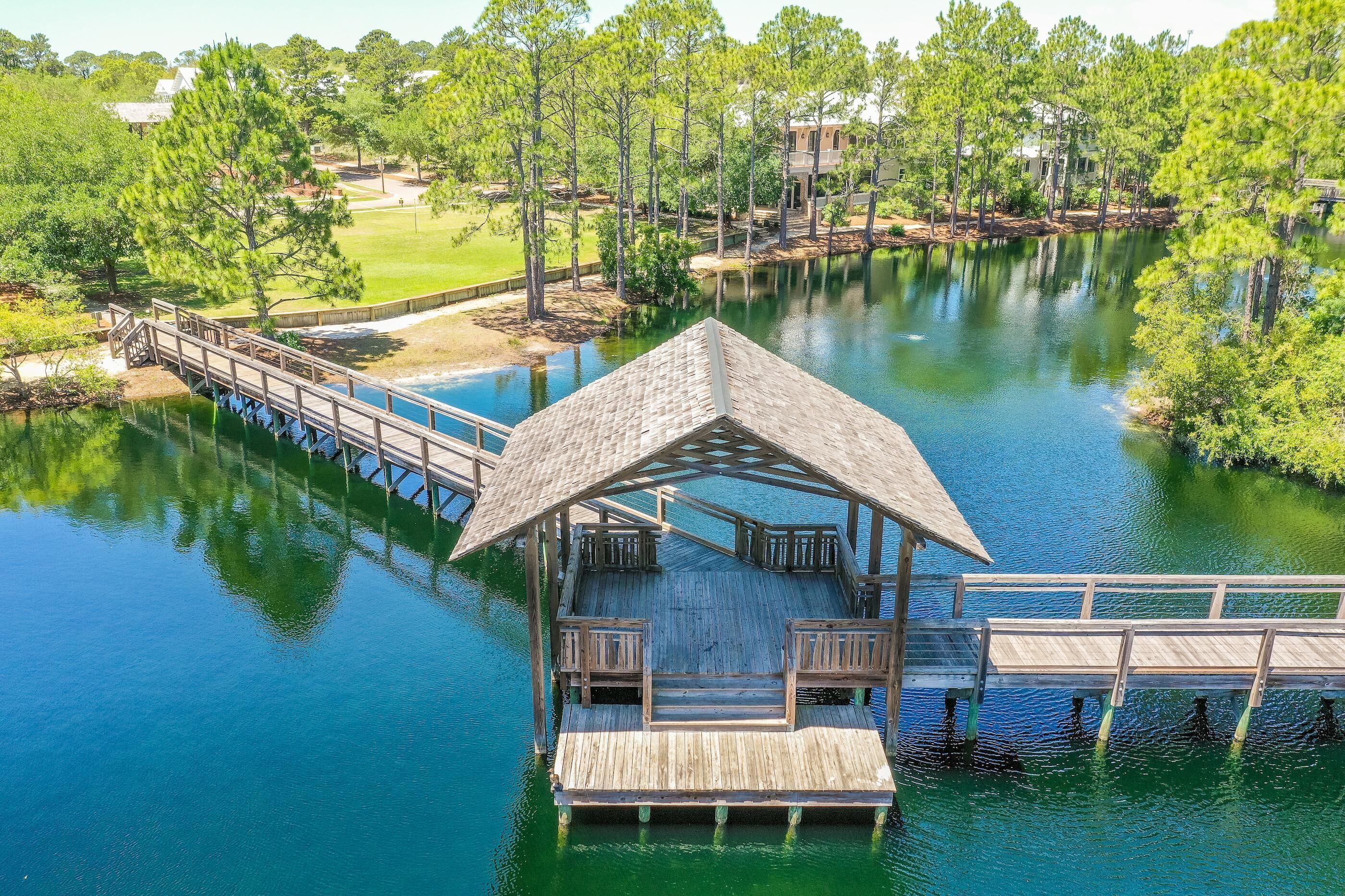 A wooden dock extending over a calm lake, surrounded by trees and a small pavilion with benches.