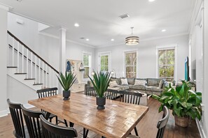 A spacious living area with a wooden dining table, comfortable seating, a staircase, and large windows. - The Greenhouse (Santa Rosa Beach)