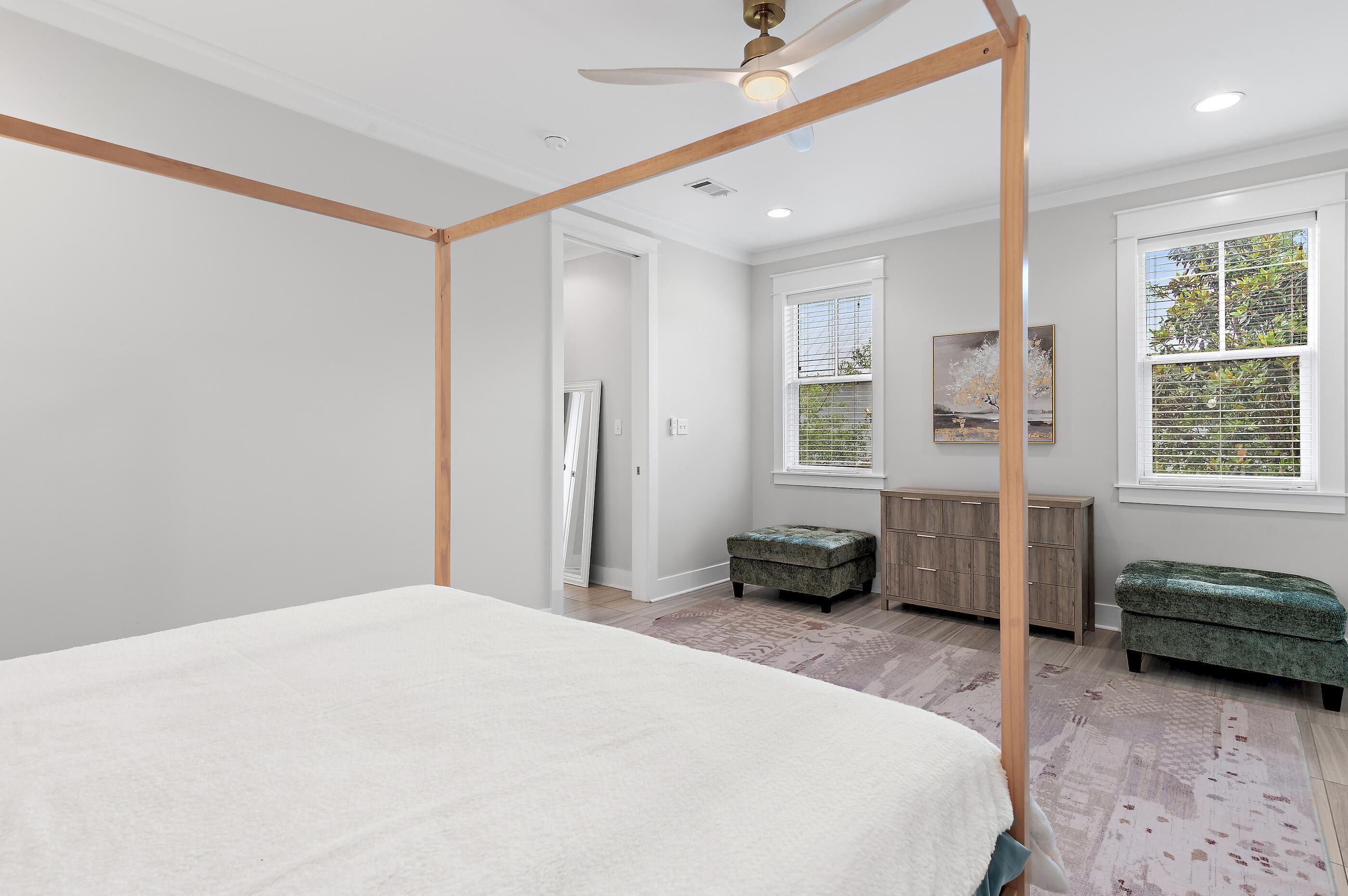 A bedroom with a four-poster bed, a wooden ceiling fan, a large window with blinds, a green upholstered bench, and a wooden cabinet.