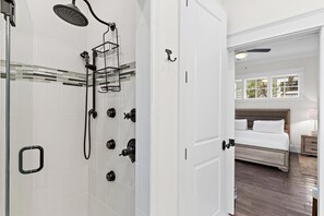 Uma casa de banho moderna com cabine de duche em vidro, porta branca e quarto visível com cama e mesa de cabeceira.