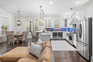 A spacious kitchen with white cabinetry, a stainless steel refrigerator, and a dining area with wicker chairs and a brown sofa. - Jaybird Bungalow (Santa Rosa Beach)