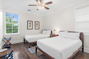 Quarto com duas camas, um ventilador de teto, janela com vista para vegetação e duas imagens emolduradas na parede.