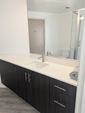 A modern bathroom with a large mirror, a sink, dark wood cabinets, and a glass shower enclosure. - Beautiful luxury high-rise condo with views of downtown Los Angeles in Koreatown (Vernon)