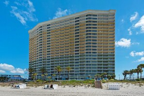 Ein Hochhaus-Hotel mit mehreren Balkonen, gelegen an einem Strand mit Palmen und Sonnenschirmen.