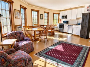 A spacious living room with a patterned sofa, a dining table with chairs, a kitchen area with white cabinets and appliances, and large windows allowing natural light.