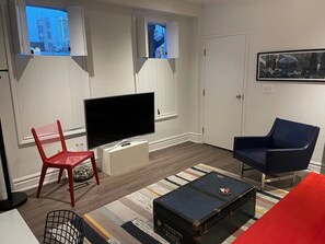 Una sala de estar con un televisor de pantalla plana, una silla roja, un sillón azul, un tapete estampado y un caja de almacenamiento negra.