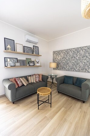 Una sala de estar con dos sofás grises, una mesita de mimbre, obras de arte enmarcadas en estanterías, un aire acondicionado instalado en la pared y un tapiz decorativo.