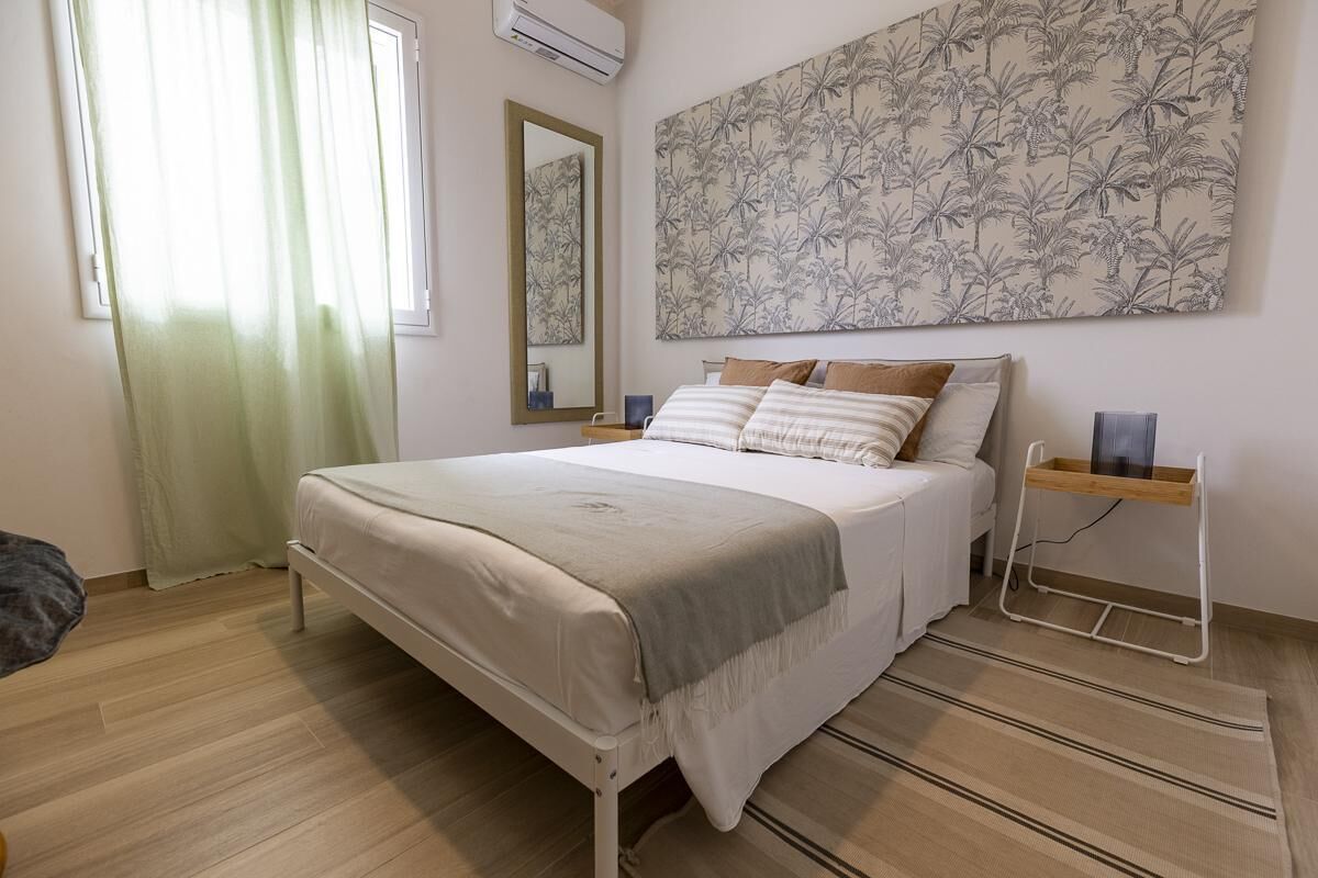 A bedroom with a bed, a bedside table, a mirror, a wall-mounted artwork, and a window with curtains.