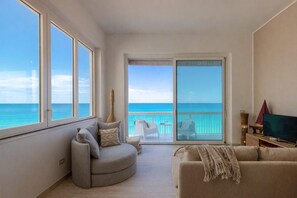 Ein Wohnzimmer mit einer gebogenen grauen Couch, einer beigen Couch und einem Fernseher auf einem Holzständer. Ein großes Fenster bietet Blick auf den Ozean.