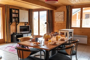 Ein gemütlicher Essbereich mit einem für das Frühstück gedeckten Tisch, darunter Croissants und Saft, und einem Fernseher im Hintergrund.