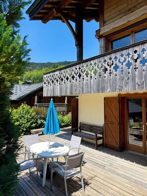 Eine Holzterrasse mit einem runden Tisch, Stühlen und einer Bank, sowie einem blauen Sonnenschirm.