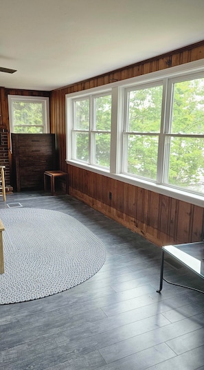 A room with a large window, wooden walls, a dark floor, and a round rug. - 3-bedroom cottage on Forest Lake in Windham (Windham)