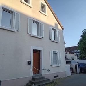 Un bâtiment à deux étages avec des murs extérieurs blancs et des volets, une porte d’entrée et un escalier menant à l’entrée.