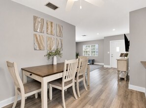 A dining area with a wooden table and chairs, a vase with greenery, and a window with blinds. - Coastal Naples Escape Private Pool, Sleeps 12, Vanderbilt Beach 5 mins away (Naples)