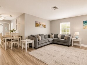 A living room with a sectional sofa, a dining table with chairs, a small side table, a lamp, and artwork on the walls.
